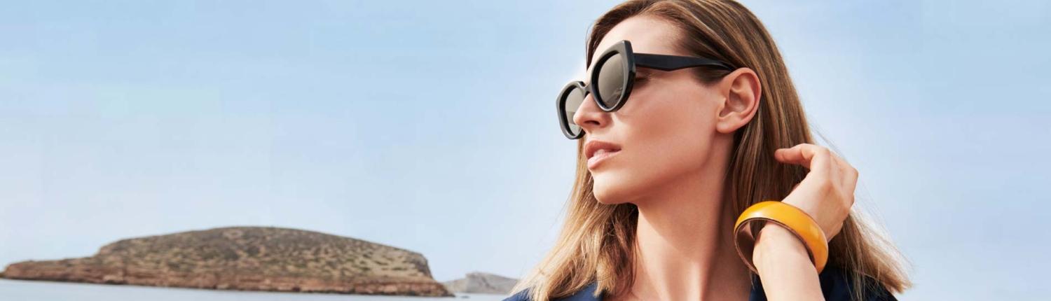 Soleil Sonnenpflege Eine Frau mit Sonnenbrille und gelbem Armreif blickt vor einer sonnigen Küstenlandschaft mit einer kleinen Insel im Hintergrund in die Ferne.