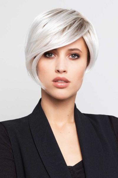 Eine Frau mit einer modernen, platinblonden Perücke im Kurzhaarschnitt und einem eleganten schwarzen Blazer vor einem hellen Hintergrund.