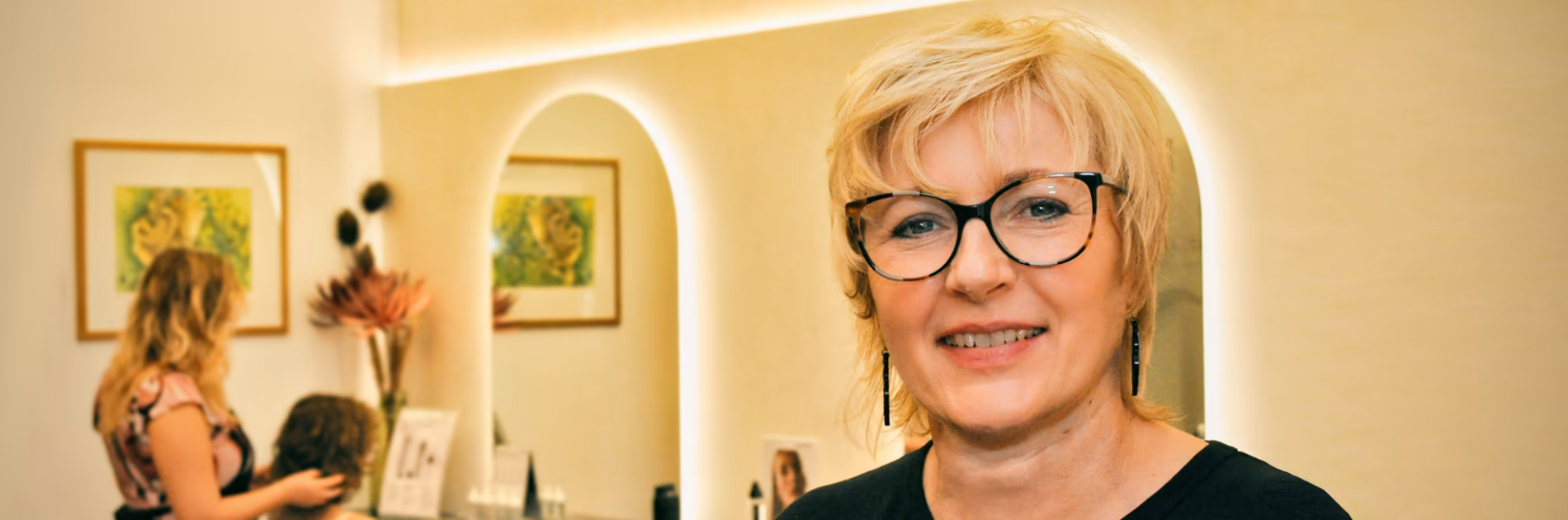 Die Friseurmeisterin Birgit Gmeiner lächelt freundlich in Ihrem hellen, modern gestalteten Friseursalon, in Bad Ischl, mit beleuchteten Spiegeln im Hintergrund.