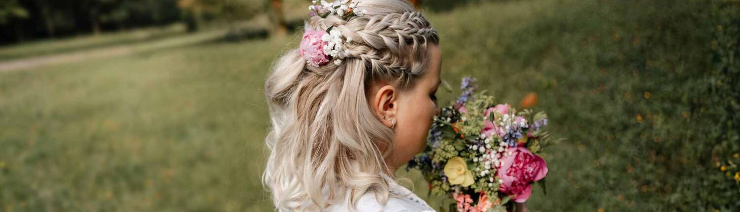 Gmeiner romantische Hochzeitsfrisur 07 2026 Eine Braut mit blondem Haar, das zu einem geflochtenen Zopf mit Blumen geschmückt ist, hält einen bunten Blumenstrauß in einer grünen Wiese.
