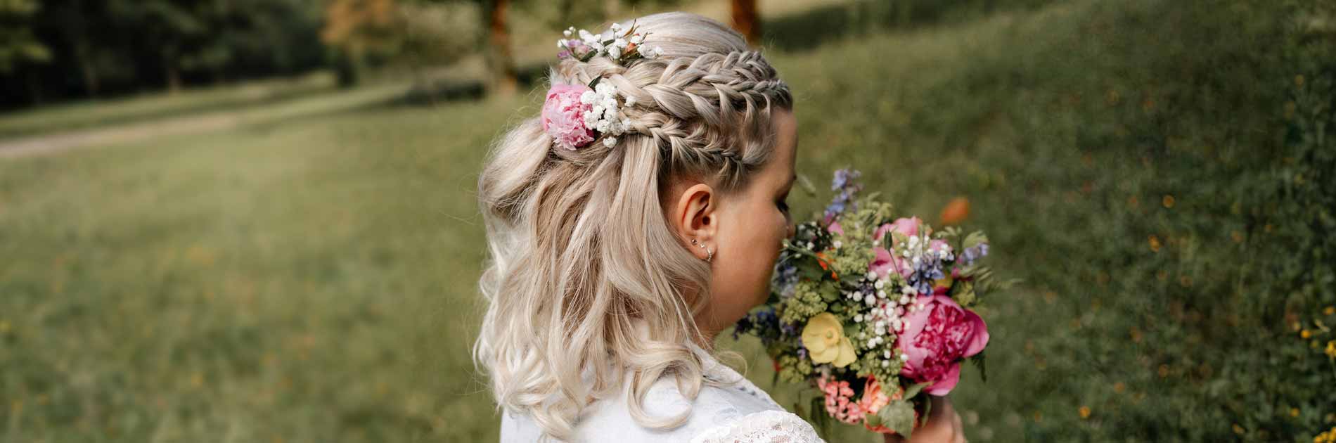Gmeiner romantische Hochzeitsfrisur 07 2026 Eine Braut mit blondem Haar, das zu einem geflochtenen Zopf mit Blumen geschmückt ist, hält einen bunten Blumenstrauß in einer grünen Wiese.