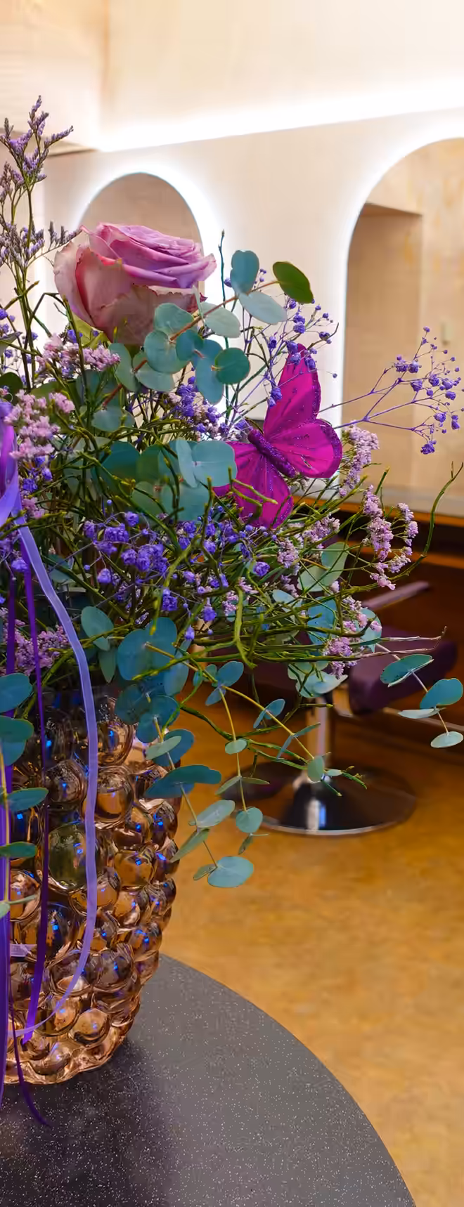 Ein Blumenstrauß mit einer lila Rose, Eukalyptus und einem künstlichen Schmetterling in einer strukturierten Glasvase auf einem dunklen Tisch.