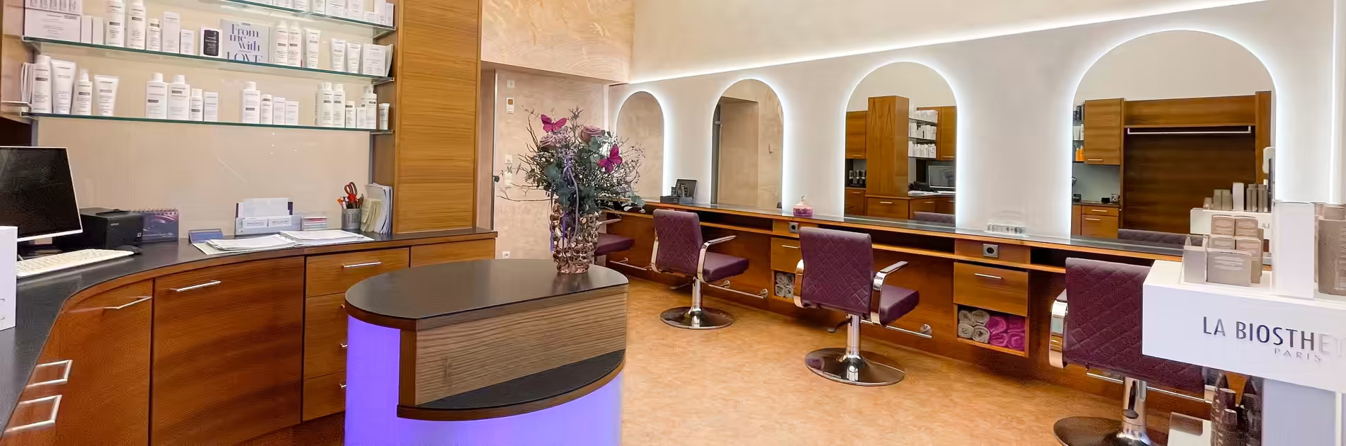 Moderner Friseursalon Birgit Gmeiner mit eleganten lila Stühlen, beleuchteten Spiegeln und hochwertigen Pflegeprodukten in einem warmen Ambiente.
