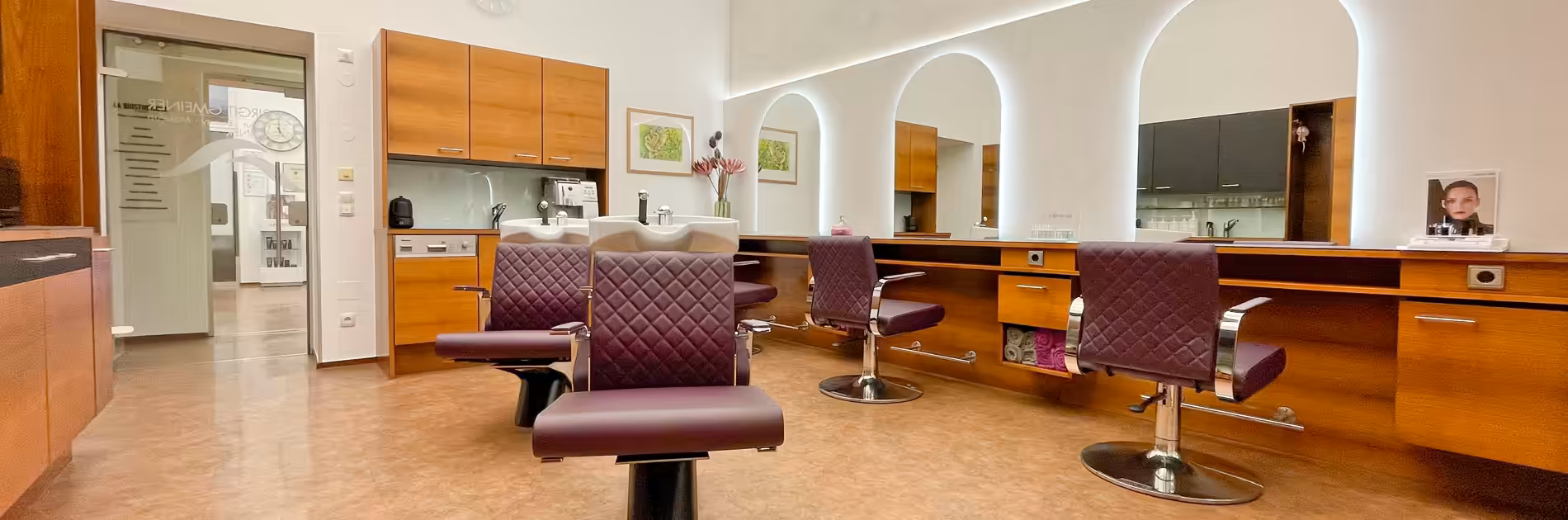 Friseursalon Birgit Gmeiner Raum zwei Ein moderner Friseursalon mit hellen Wänden, Holzelementen, drei violetten Bedienplätzen und großen, beleuchteten Spiegeln in einem hellen Raum.