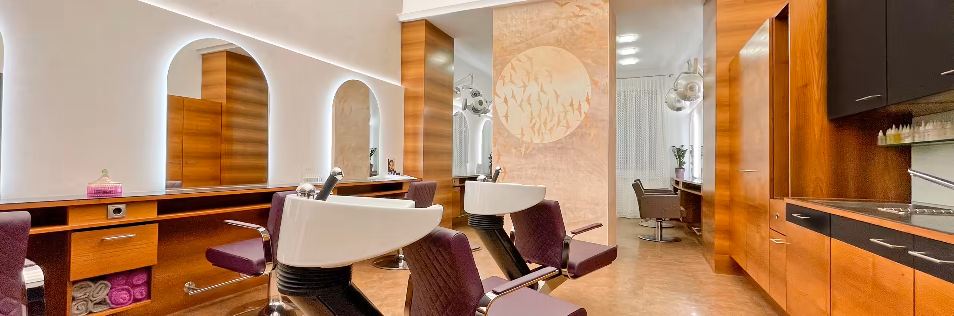 Friseursalon Birgit Gmeiner Raum zwei Modern eingerichteter Friseursalon Birgit Gmeiner mit hellen Spiegeln, bequemen lila Sesseln und professionellen Waschbecken in warmer Holzoptik.