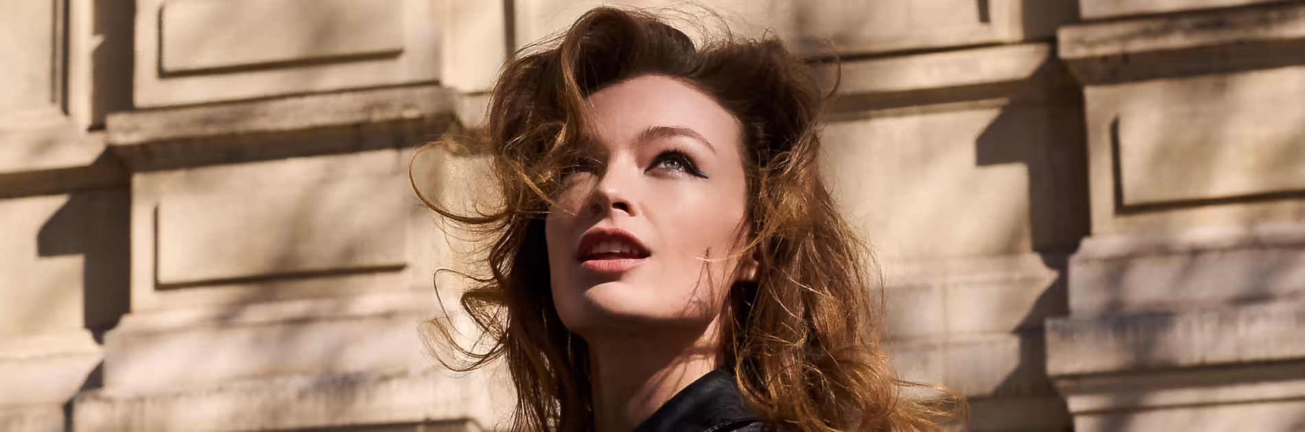 Eine Frau mit voluminösem Haar und elegantem Make-up blickt nach oben, passend zum Make-Up Trend Awakened vor einer klassischen Fassade.